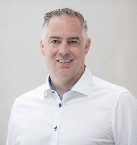 A man with short gray hair wearing a white button-up shirt is smiling and standing against a plain light background.