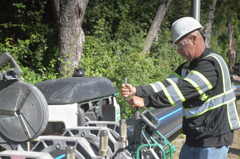 A construction worker in a hard hat and safety gear operates a machine for joining pipes outdoors. He turns a handle near a long blue pipe, set against a backdrop of trees and greenery.