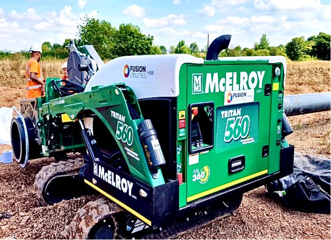 A green McElroy Tritan 560 machine is on a construction site with a man standing beside it. The machine is mounted on tracks, surrounded by gravel and greenery, under a clear blue sky.
