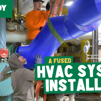 Several workers install a large blue HVAC pipe indoors. Text reads “McElroy” and “A Fused HVAC System Install!!” in bold green and white letters. Tools and machinery surround the workers.