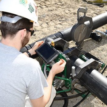 A person wearing a helmet and sunglasses uses a handheld device to inspect large black pipes on a sandy surface. The pipes are connected to machinery with various gauges and cables.