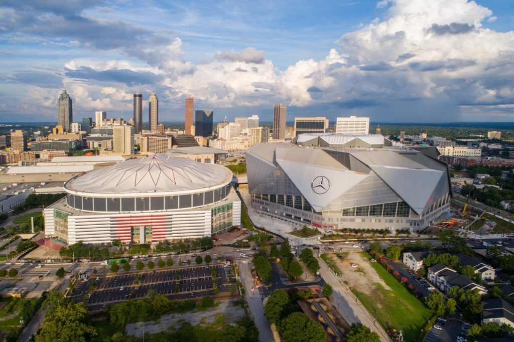 Aerial view of Atlantas downtown skyline featuring two stadiums: the Georgia Dome with its circular design and the angular, modern Mercedes-Benz Stadium. Skyscrapers, clouds, and McElroy properties punctuate the vibrant background.