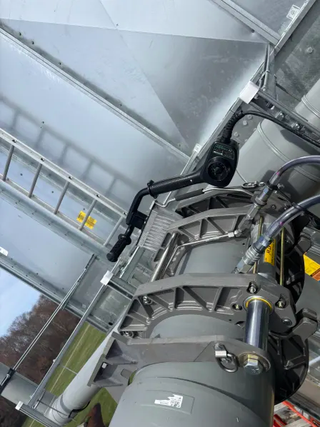 Close-up of industrial machinery with metal pipes, hydraulic components, and wiring. A metal ladder and large metallic wall structure are visible in the background. The photo is taken outdoors at an angle.