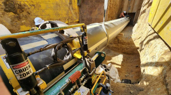 Workers install a large metal pipe in a narrow trench with yellow safety barriers. Machinery with “Danger: Crush Zone” signs is positioned near the pipe. The area appears to be part of a construction site.