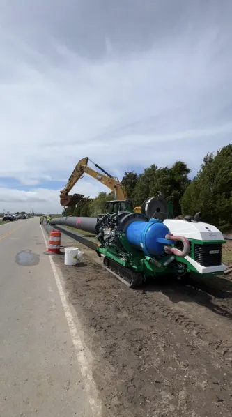 A large construction machine working on the side of a road, handling a long pipe. Orange traffic barrels line the road, and trees are visible in the background under a partly cloudy sky.