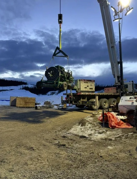 A crane lifts a large green machine at a snowy outdoor worksite during dusk. Utility trucks and construction equipment are parked nearby under cloudy skies, with snow-covered ground in the background.