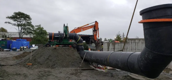 A construction site with large black plastic pipes being installed, a pile of gravel, an orange excavator, and workers in safety gear under a cloudy sky. Trees and construction materials are visible in the background.