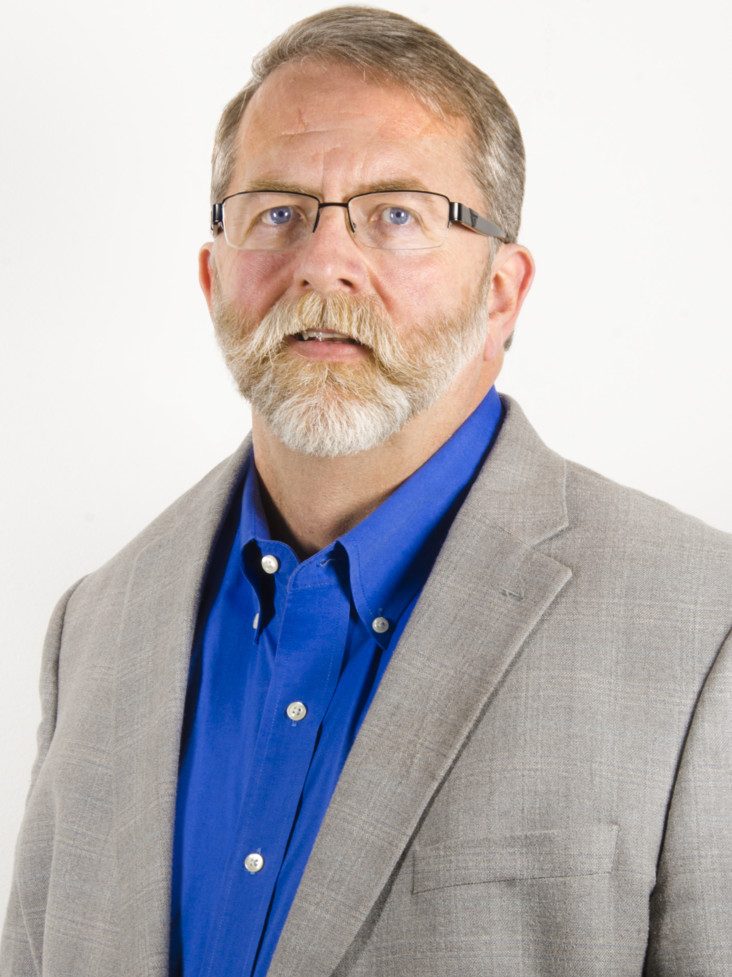 A middle-aged man with glasses, a gray mustache, and beard, wearing a blue dress shirt and a light gray blazer, stands in front of a plain white background.