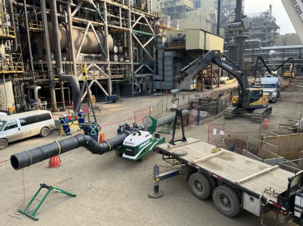 A construction site with workers, excavators, a large industrial building, a truck, and various pipes and equipment arranged on a dirt lot. Orange cones and fencing mark off work areas.