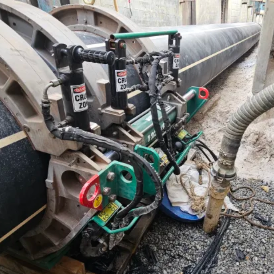 Large industrial pipe fusion machine joining two black plastic pipes at a construction site, surrounded by concrete walls and gravel, with hoses and cables connected to the equipment.