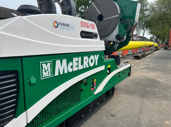 A green and white McElroy TracStar 900 machine from Fusion Utilities is parked on a road, next to yellow pipes and construction barriers, with trees and buildings in the background.