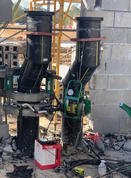 Two large black pipes are being fused at a construction site. Welding equipment is attached to the pipes, surrounded by tools, cables, and a red box. Unfinished cinder block walls and scaffolding are visible in the background.