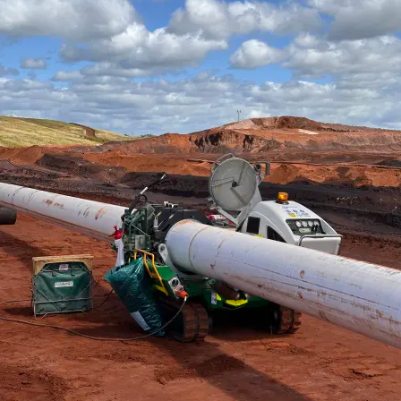 A robot with green bags and a satellite dish is stationed on a large white pipeline in a red dirt landscape under a partly cloudy sky.