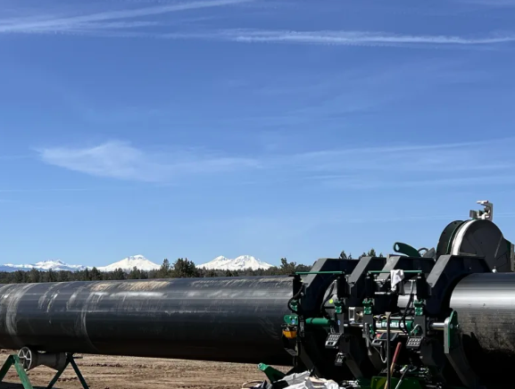 A large metal pipeline stretches across a dirt field under a blue sky, with snow-capped mountains visible in the distant background. Machinery is attached to the pipeline in the foreground.