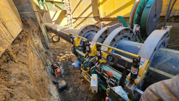 Large machinery fuses two sections of black pipe in a deep trench with yellow retaining walls. Tools and equipment are scattered nearby, and a ladder leans against the trench wall.
