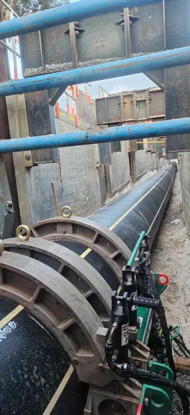 A large black pipeline is being laid in a trench supported by metal and wooden bracing. Construction equipment and sand surround the pipe, and safety barriers and fencing are visible above the trench.