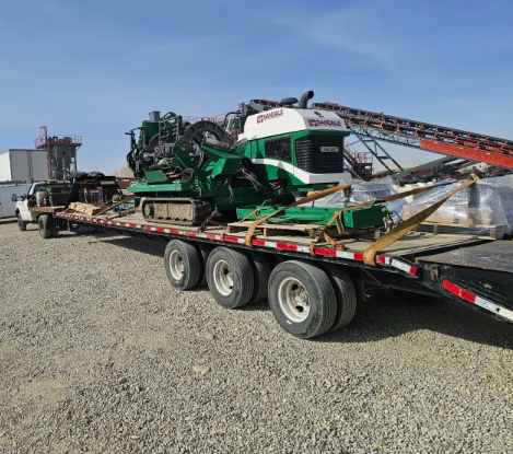 A large green industrial machine is secured on a flatbed trailer with wooden braces. The trailer is parked on gravel, next to a pickup truck, with industrial equipment and conveyor belts in the background.
