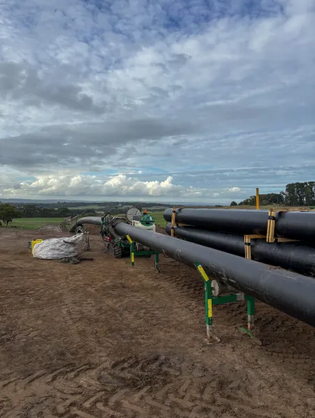 Large black pipelines are laid out on a dirt construction site with green supports, under a cloudy sky. A worker in safety gear is near the pipes, and farmland is visible in the background.