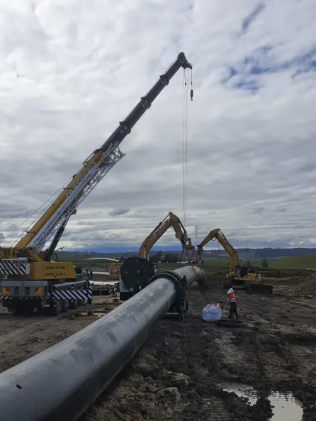 Two cranes and an excavator are lifting and positioning a large pipeline segment on a muddy construction site, with workers in safety gear nearby under a cloudy sky.