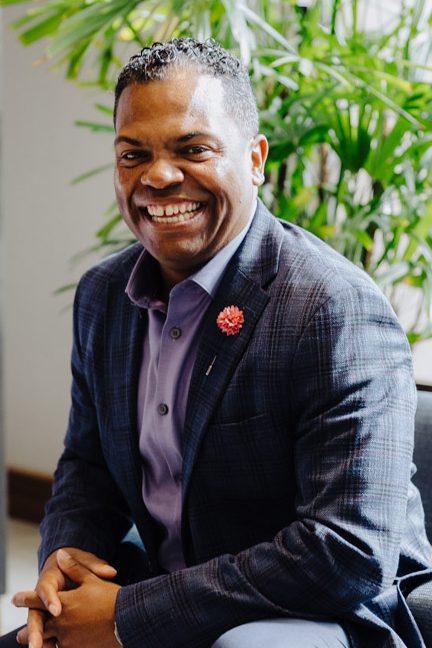 A man in a plaid jacket and purple shirt sits smiling on a dark chair, hands clasped. A green plant and modern decor are in the background, creating a bright, welcoming atmosphere.
