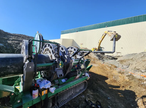 A large green pipe fusion machine operates on a construction site with dirt and rocks, while a yellow excavator lifts a pipe near a beige industrial building under a clear blue sky.