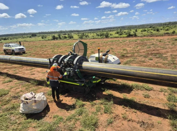 A worker in a high-visibility vest operates a large machine to join sections of black pipeline in a rural field, with a utility vehicle and open landscape in the background under a partly cloudy sky.