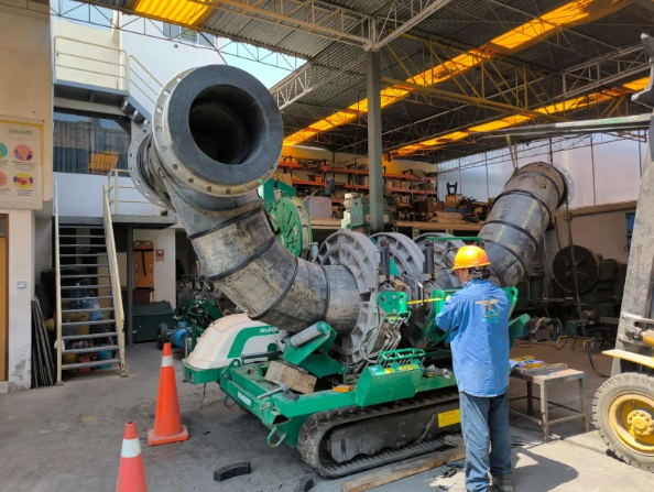 A worker in a blue shirt and yellow helmet operates a large green industrial machine with thick, curved black pipes inside a workshop with shelves, tools, and orange safety cones.