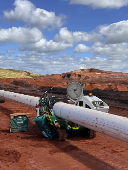 A robotic device on tracks inspects a large white pipeline laid across reddish earth at an industrial site, with cloudy blue sky and layered hills in the background.