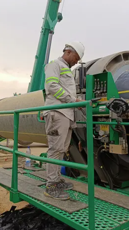 A worker in a white helmet and reflective safety gear operates machinery on a green metal platform at a construction site, with large pipes and equipment in the background.