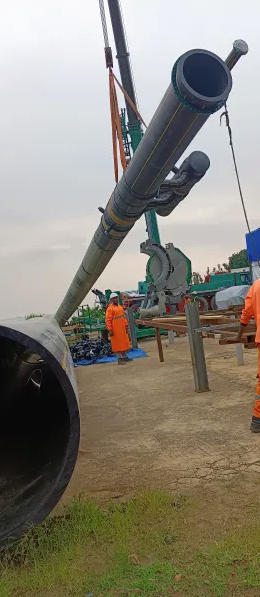 Workers in orange safety clothing manage the installation of a large pipeline using a crane at an industrial outdoor site, with cloudy skies overhead and equipment visible in the background.