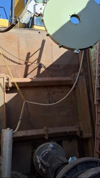 A large circular apparatus with cables is suspended above industrial equipment in a rust-colored metal enclosure, with pipes and wires running along the walls. Sunlight casts shadows onto the surfaces.