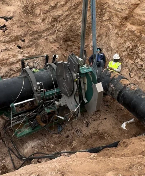 Two construction workers in safety vests are working in a dirt trench with large black pipes, using heavy machinery to join or repair the pipeline.