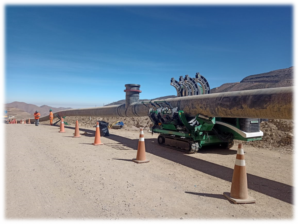A large industrial pipeline is being worked on in a desert area. Traffic cones line the dirt road, while workers in orange safety gear and a green construction machine are positioned alongside the pipe. Mountains are visible in the background.