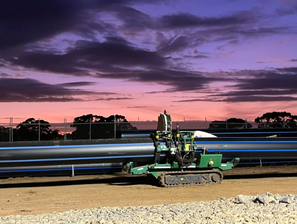 A green tracked machine sits on dirt in front of large pipes at a construction site, with a colorful sunset sky and silhouetted trees in the background.