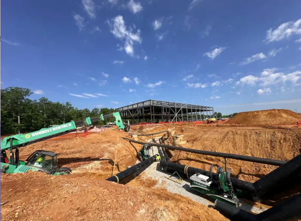 Construction site with large steel building frame in background, heavy machinery, green equipment, and large pipes being installed in a dirt excavation under a blue sky with scattered clouds.