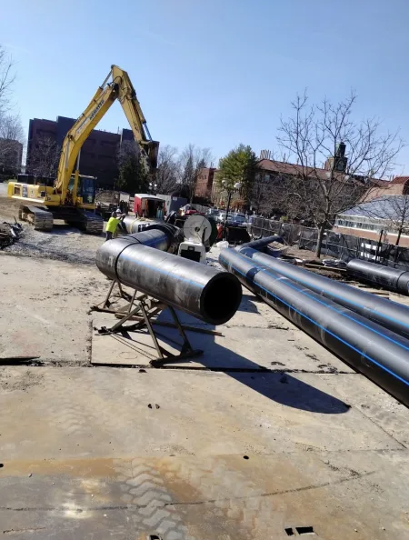 Large steel pipes are stacked at a construction site, with an excavator and several workers in safety vests nearby. Trees and buildings are visible in the background under a clear sky.