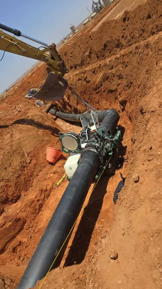 A large black pipeline is being installed in a deep trench in reddish soil, with heavy machinery and equipment visible, including an excavator and a pipe fusion machine. Construction materials are scattered around the site.