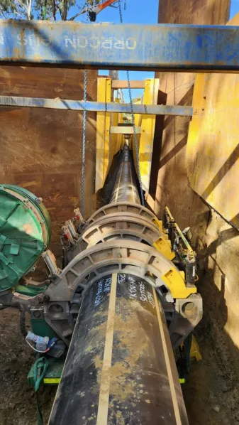A large black pipeline is being welded and joined using heavy equipment at a construction site, surrounded by wooden and metal supports, with dirt and machinery visible.