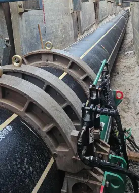 Large black pipe being welded or joined with mechanical equipment in a construction trench, surrounded by concrete walls and sandy ground.