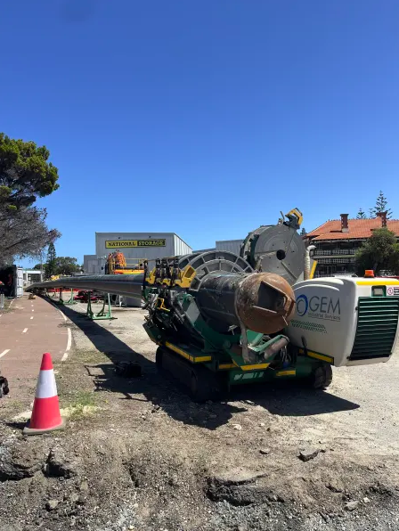Large industrial drilling machine on a roadside construction site, with an orange traffic cone nearby and a National Storage building in the background under a clear blue sky.
