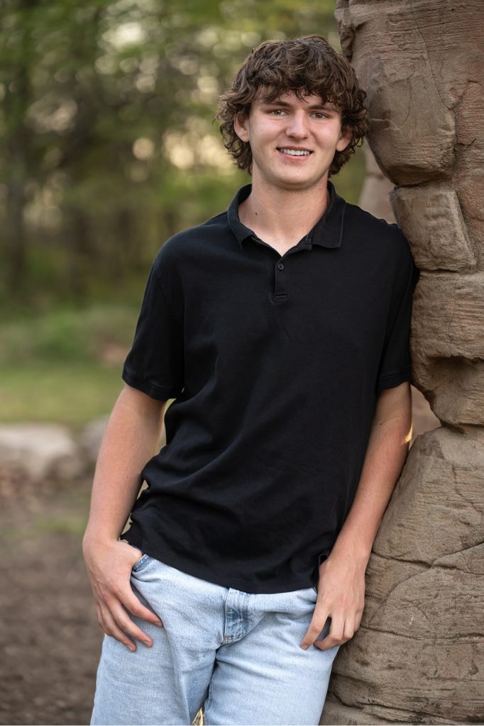 A young man with curly brown hair wearing a black polo shirt and light blue jeans leans against a large stone structure outdoors, smiling at the camera. Trees and grass are visible in the blurred background.