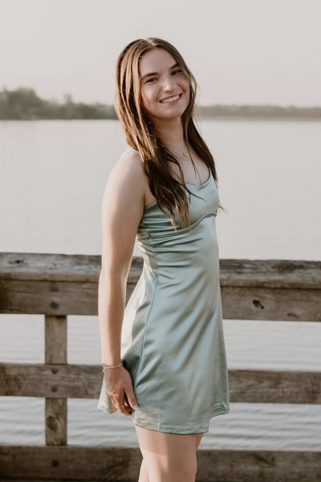 A young woman with long brown hair stands smiling on a wooden dock by a calm lake, wearing a light green satin dress. The background features water and distant trees in soft, natural light.