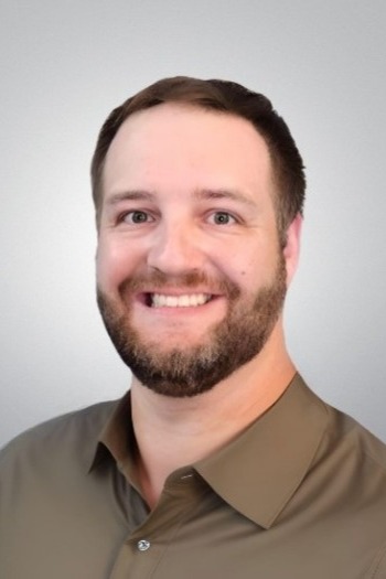 A man with short brown hair and a beard, wearing a brown collared shirt, smiling in front of a plain light gray background.