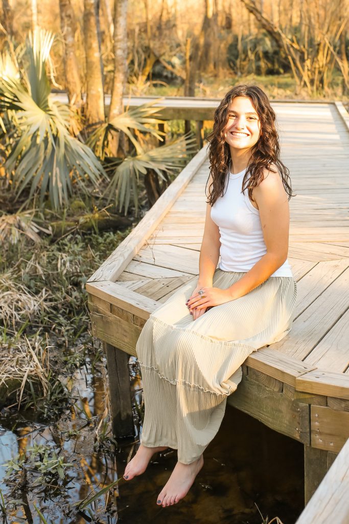 A young woman with wavy brown hair sits barefoot on a wooden boardwalk over a marsh, smiling at the camera. She wears a white sleeveless top and a long beige skirt. Sunlight filters through the trees in the background.