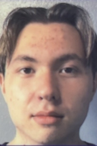 A young person with short, light brown hair parted in the middle, looking straight at the camera with a neutral expression. The background is plain and out of focus.
