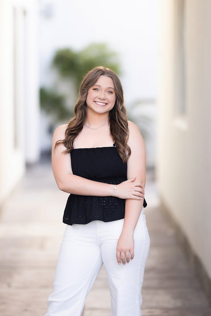 A young woman with wavy brown hair stands outdoors in a bright, narrow walkway. She is smiling, wearing a black sleeveless top and white pants, with one arm resting across her body. The background is softly blurred.