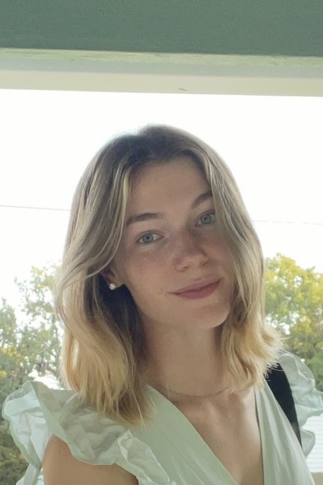 A young woman with shoulder-length blonde hair and blue eyes smiles softly at the camera. She is wearing a light-colored ruffled top and standing in front of a bright outdoor background with green trees.