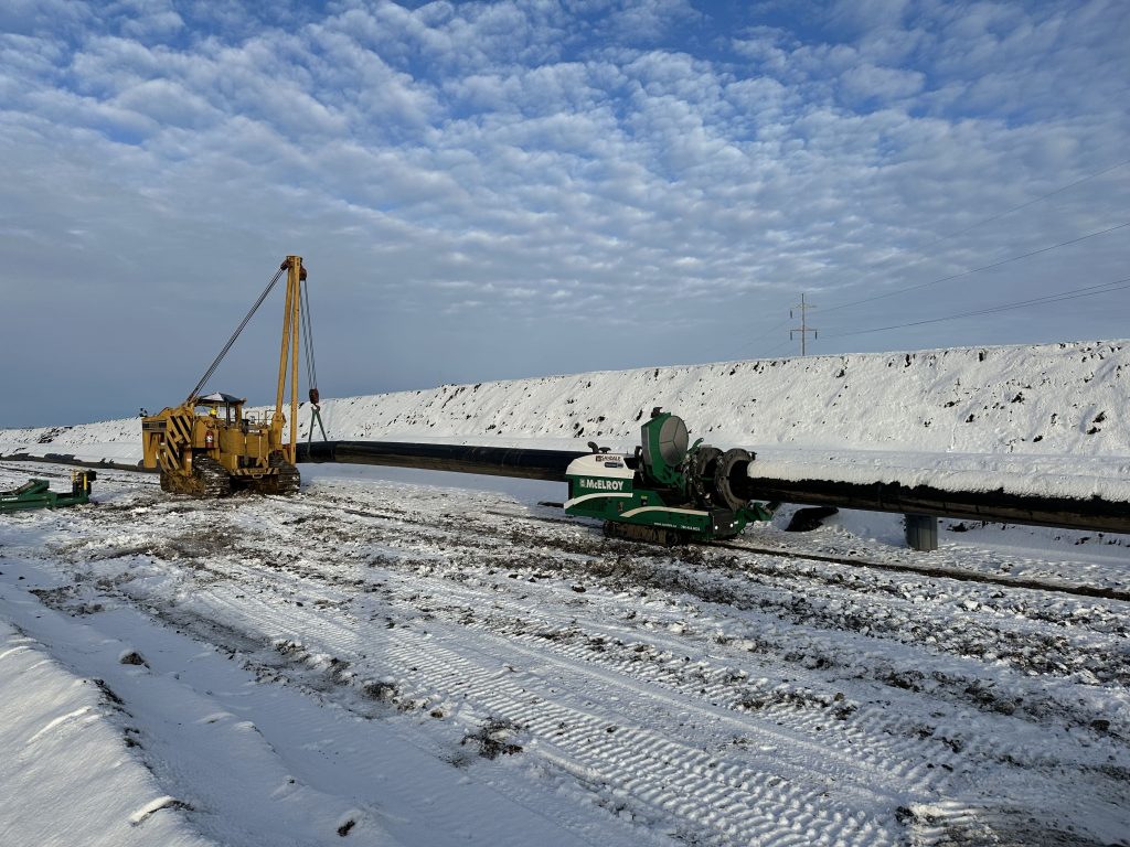 Yellow and green construction vehicles are working on installing a large pipeline across a snowy, open landscape under a partly cloudy sky. Tire tracks and machinery marks are visible in the snow.