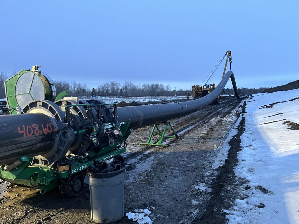 A large black pipeline is being installed outdoors on muddy ground with patches of snow, using heavy machinery. Trees and a clear sky are visible in the background.