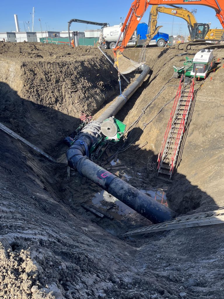 A large pipeline is being installed in a deep trench at a construction site. Excavators, equipment, and ladders surround the area. The exposed pipe is partially supported by straps and machinery.
